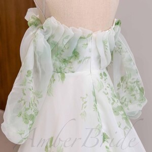 May include: A white off-the-shoulder dress with a green floral print and a ruffled neckline. The dress is made of a sheer fabric and has a flowing skirt.
