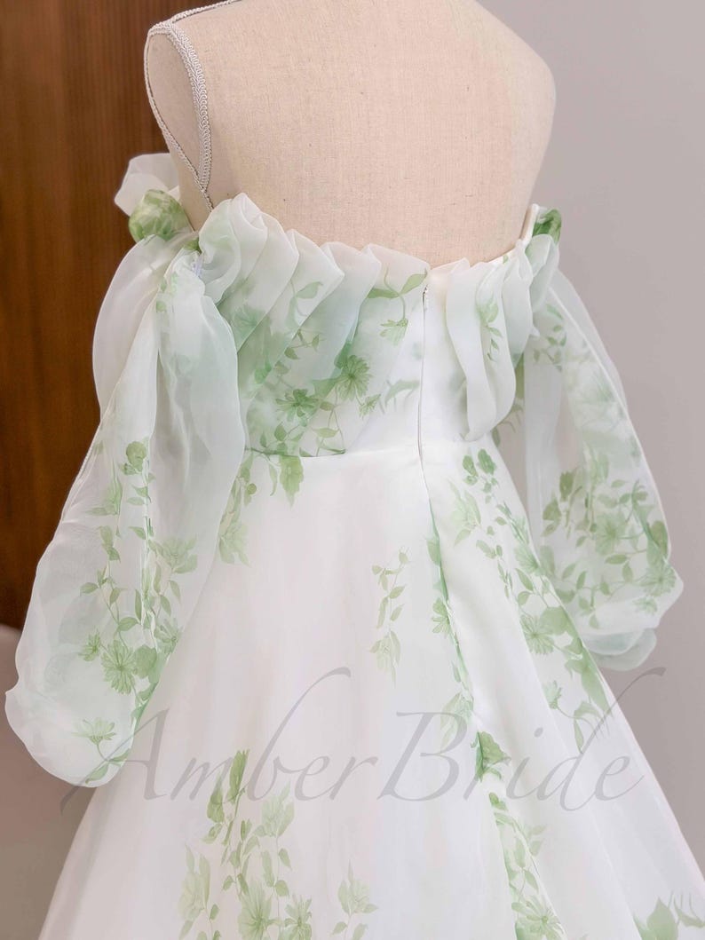 May include: A white off-the-shoulder dress with a green floral print and a ruffled neckline. The dress is made of a sheer fabric and has a flowing skirt.