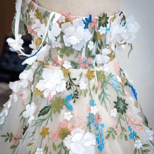 May include: A white tulle dress with a floral embroidery design. The dress features 3D flowers in white, blue, pink, and yellow. The dress has a fitted bodice and a full skirt.