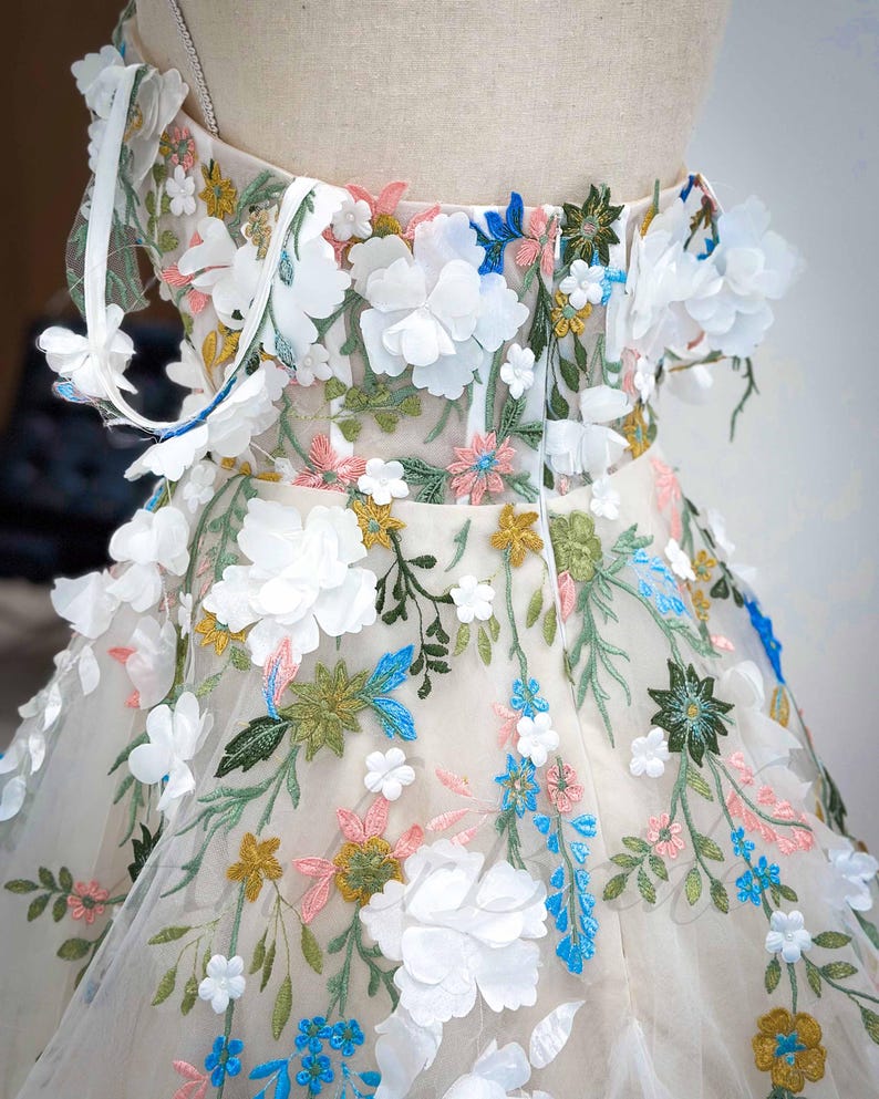 May include: A white tulle dress with a floral embroidery design. The dress features 3D flowers in white, blue, pink, and yellow. The dress has a fitted bodice and a full skirt.