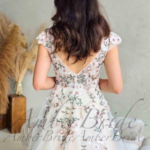 May include: A cream-colored wedding dress with floral embroidery. The dress features a V-neckline and cap sleeves, with delicate pink and green floral patterns. The dress has a tulle skirt and is suitable for a wedding or special occasion.