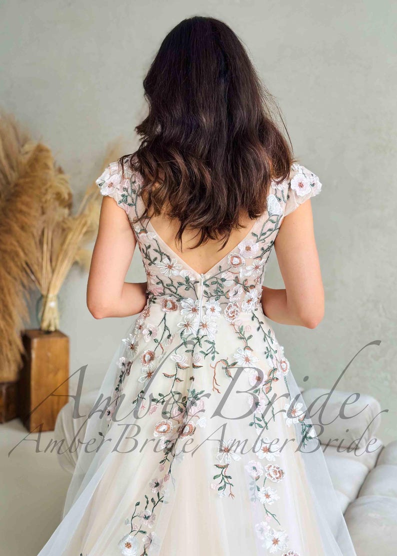 May include: A cream-colored wedding dress with floral embroidery. The dress features a V-neckline and cap sleeves, with delicate pink and green floral patterns. The dress has a tulle skirt and is suitable for a wedding or special occasion.