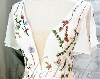 May include: White wedding dress with a plunging neckline and short, sheer sleeves. The dress is adorned with colorful floral embroidery in shades of red, green, and blue. The dress is displayed on a mannequin.