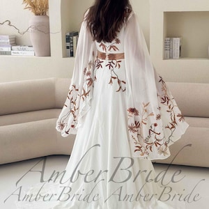 May include: A white wedding dress with floral embroidery in shades of brown and gold. The dress features a flowing skirt and a sheer cape with matching embroidery. The text "Amber Bride" is visible at the bottom of the image.