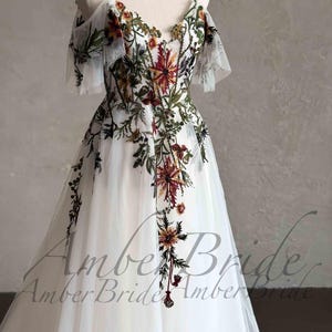 May include: A white tulle wedding dress with a floral embroidery design. The dress has a V-neckline, off-the-shoulder straps, and short sleeves. The embroidery features a variety of flowers in shades of red, orange, yellow, green, and brown.