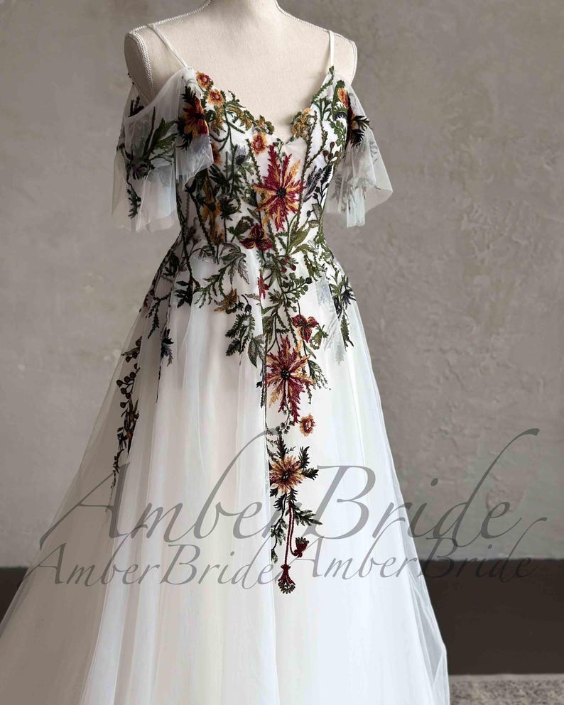 May include: A white tulle wedding dress with a floral embroidery design. The dress has a V-neckline, off-the-shoulder straps, and short sleeves. The embroidery features a variety of flowers in shades of red, orange, yellow, green, and brown.