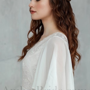 May include: A white lace wedding dress with a flowing chiffon overlay and a V-neckline. The dress has a fitted bodice and a flowing skirt. The model is wearing her hair in loose waves.