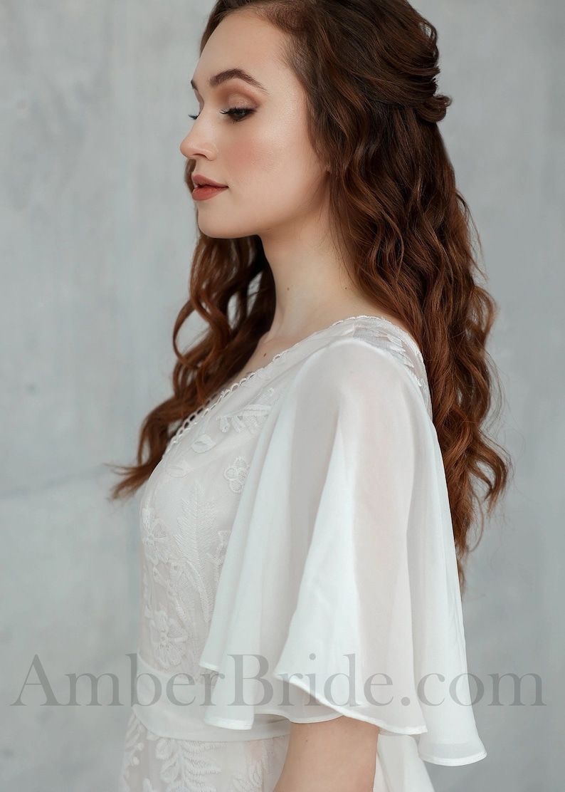 May include: A white lace wedding dress with a flowing chiffon overlay and a V-neckline. The dress has a fitted bodice and a flowing skirt. The model is wearing her hair in loose waves.