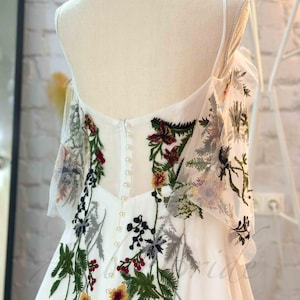 May include: White wedding dress with a floral embroidery design. The dress has a sweetheart neckline and a fitted bodice. The skirt is full and flowing. The dress is accented with pearl buttons down the back.