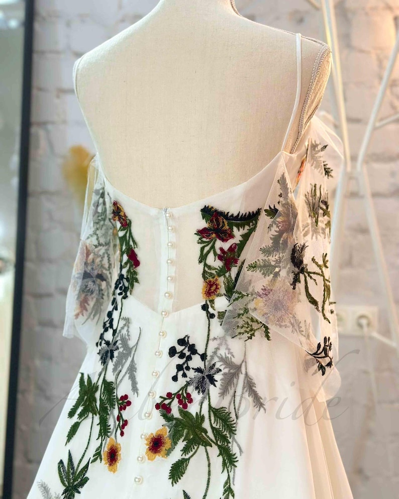 May include: White wedding dress with a floral embroidery design. The dress has a sweetheart neckline and a fitted bodice. The skirt is full and flowing. The dress is accented with pearl buttons down the back.