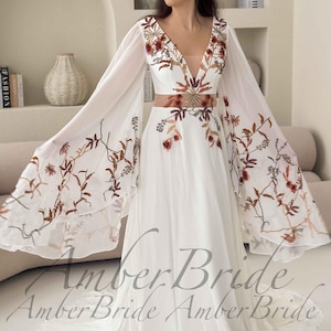 May include: White wedding dress with a plunging neckline and long, flowing sleeves. The dress features floral embroidery in shades of brown and red. The waistline is accentuated with a matching embroidered belt. The dress is from Amber Bride.