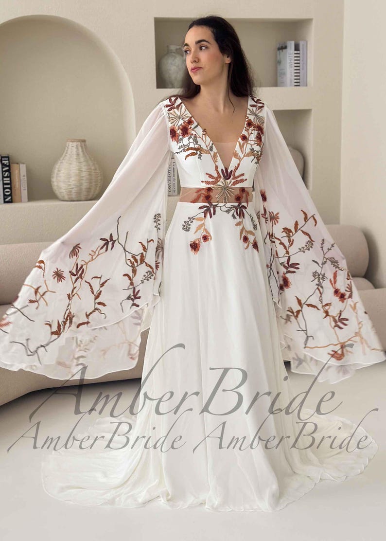 May include: White wedding dress with a plunging neckline and long, flowing sleeves. The dress features floral embroidery in shades of brown and red. The waistline is accentuated with a matching embroidered belt. The dress is from Amber Bride.