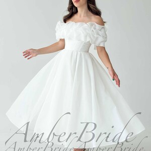 May include: A white off-the-shoulder wedding dress with a ruffled neckline and short sleeves. The dress has a fitted bodice and a flowing skirt. The image shows the dress in a studio setting, with the model's arms outstretched.
