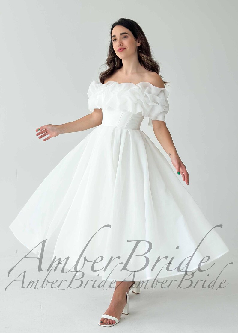 May include: A white off-the-shoulder wedding dress with a ruffled neckline and short sleeves. The dress has a fitted bodice and a flowing skirt. The image shows the dress in a studio setting, with the model's arms outstretched.