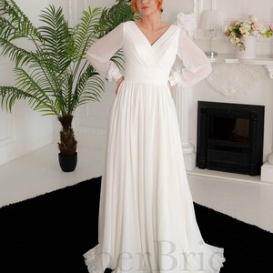 May include: A white chiffon wedding dress with long sleeves and a V-neckline. The dress has a fitted bodice and a flowing skirt.