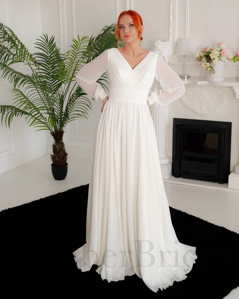 May include: A white chiffon wedding dress with long sleeves and a V-neckline. The dress has a fitted bodice and a flowing skirt.