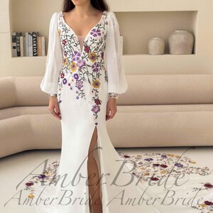 May include: A white wedding dress with a floral embroidered bodice and a high slit. The dress features long, sheer sleeves and a train with matching floral embroidery. The text "Amber Bride" is visible at the bottom of the dress.