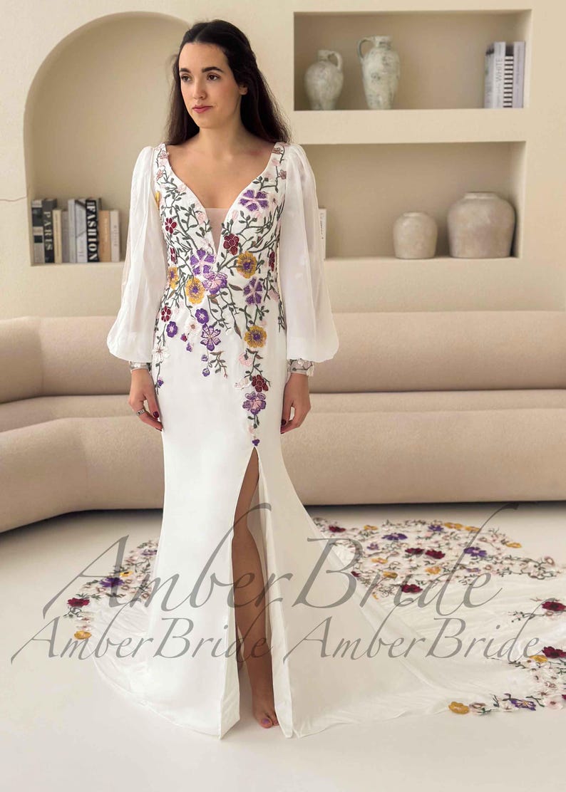May include: A white wedding dress with a floral embroidered bodice and a high slit. The dress features long, sheer sleeves and a train with matching floral embroidery. The text "Amber Bride" is visible at the bottom of the dress.