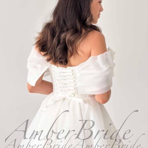 May include: A white off-the-shoulder wedding dress with a corset back and a flowing skirt. The dress features sheer, ruffled sleeves and a bow at the waist. The text "Amber Bride" is visible on the lower part of the dress.