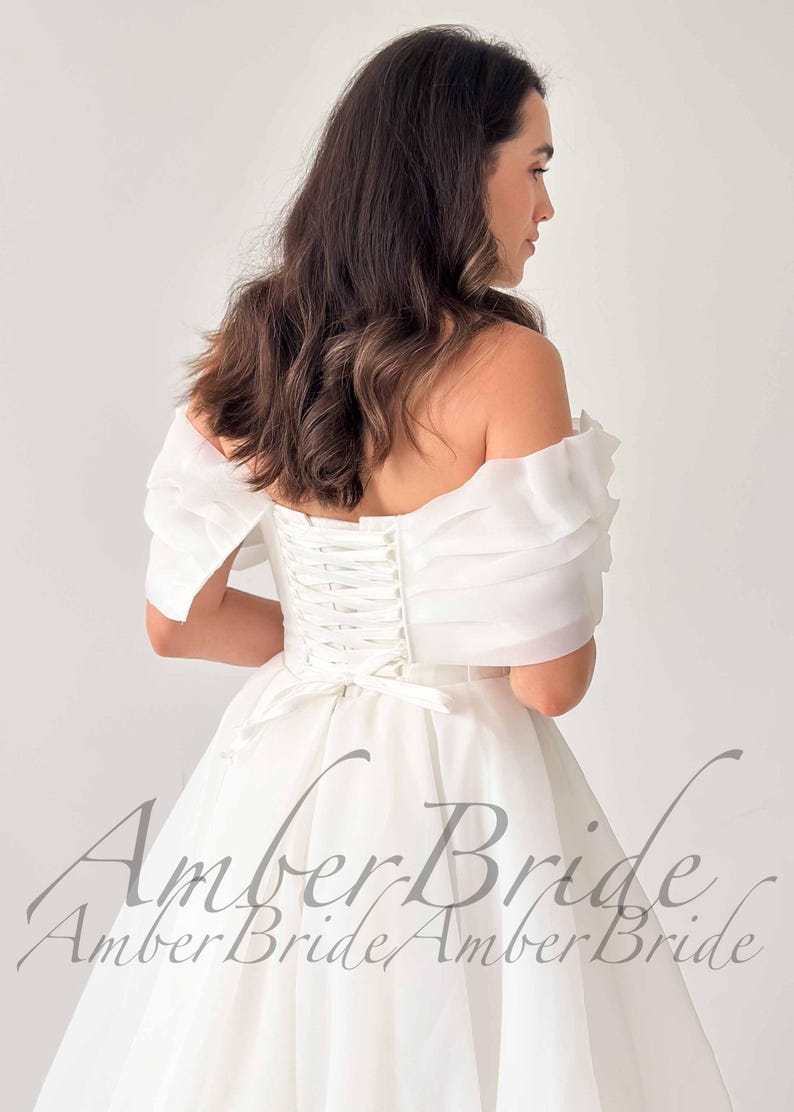 May include: A white off-the-shoulder wedding dress with a corset back and a flowing skirt. The dress features sheer, ruffled sleeves and a bow at the waist. The text "Amber Bride" is visible on the lower part of the dress.