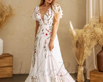 May include: A white wedding dress with a plunging neckline and short, flutter sleeves. The dress is adorned with delicate floral embroidery in shades of red, green, and yellow. The skirt flows to the floor, creating an elegant silhouette.