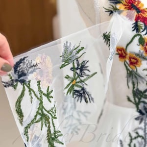 May include: White tulle fabric with embroidered floral designs in shades of green, blue, yellow, and red. The flowers are delicate and intricate, and the fabric is soft and sheer.
