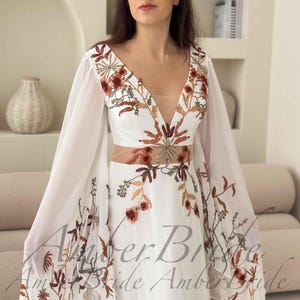 May include: A white wedding dress with a plunging neckline and sheer sleeves. The dress features intricate floral embroidery in shades of brown and orange. The text "Amber Bride" is visible on the dress.