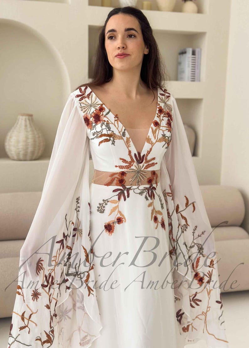 May include: A white wedding dress with a plunging neckline and sheer sleeves. The dress features intricate floral embroidery in shades of brown and orange. The text "Amber Bride" is visible on the dress.