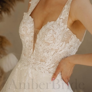 Könnte beinhalten: Weißes Brautkleid mit tiefem V-Ausschnitt und aufwendigen floralen Spitzendetails. Das Kleid hat ein figurbetontes Oberteil und einen fließenden Rock. Der Text "Amber Bride" ist auf dem Rock zu sehen.
