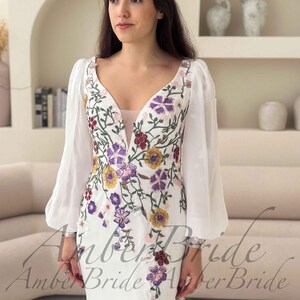 May include: White wedding dress with floral embroidery in purple, yellow, and red. The dress features a V-neckline, sheer sleeves, and a thigh-high slit. The text "Amber Bride" is visible on the dress.