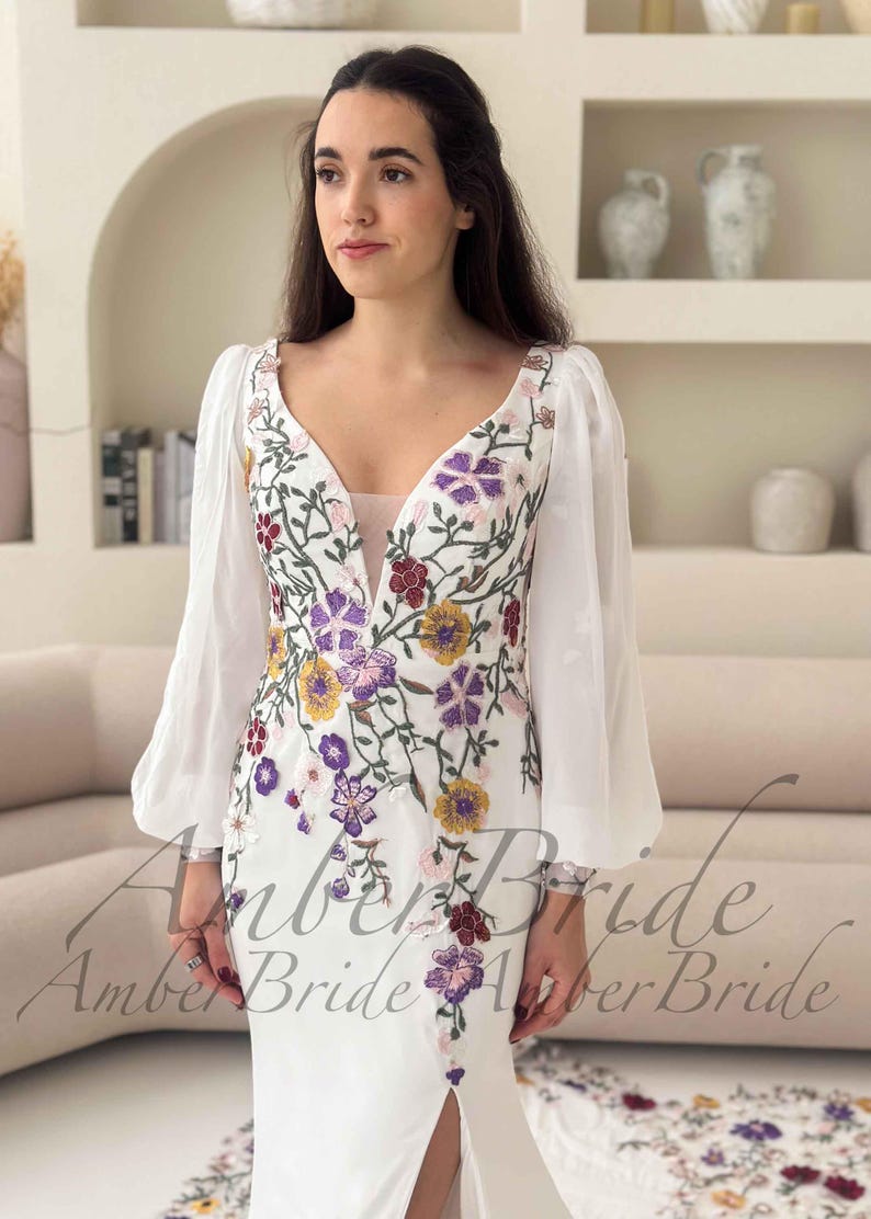May include: White wedding dress with floral embroidery in purple, yellow, and red. The dress features a V-neckline, sheer sleeves, and a thigh-high slit. The text "Amber Bride" is visible on the dress.