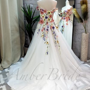 May include: A white off-the-shoulder tulle gown with colorful floral embroidery. The gown has a full skirt and a fitted bodice. The embroidery features a variety of flowers, including roses, daisies, and sunflowers. The gown is perfect for a special occasion, such as a wedding or prom.