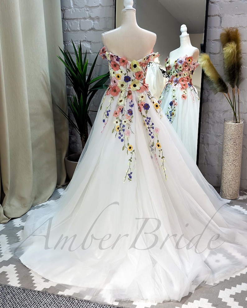 May include: A white off-the-shoulder tulle gown with colorful floral embroidery. The gown has a full skirt and a fitted bodice. The embroidery features a variety of flowers, including roses, daisies, and sunflowers. The gown is perfect for a special occasion, such as a wedding or prom.