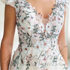 May include: Ivory wedding dress with a V-neckline and cap sleeves. The dress is adorned with delicate floral embroidery in white, pink, and brown, with green leaves. The bodice and sleeves are sheer, creating a romantic look.