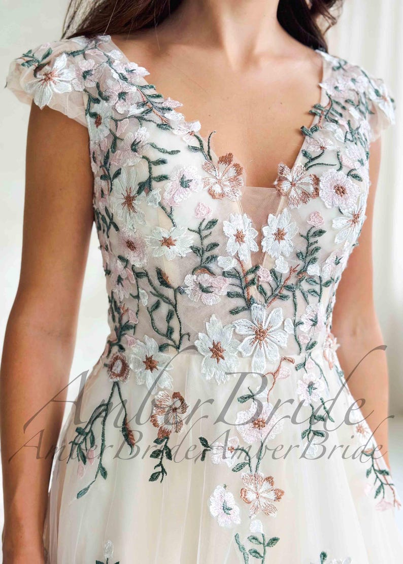 May include: Ivory wedding dress with a V-neckline and cap sleeves. The dress is adorned with delicate floral embroidery in white, pink, and brown, with green leaves. The bodice and sleeves are sheer, creating a romantic look.