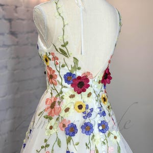 May include: A white tulle dress with colorful embroidered flowers. The dress has a V-neckline and a sleeveless design. The back of the dress features a row of buttons.