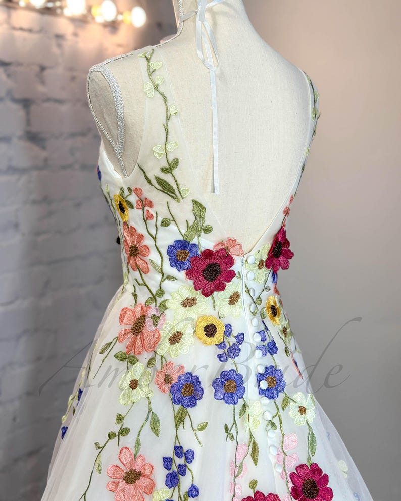May include: A white tulle dress with colorful embroidered flowers. The dress has a V-neckline and a sleeveless design. The back of the dress features a row of buttons.