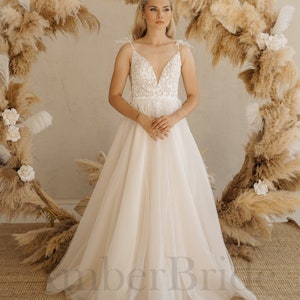 May include: A white wedding dress with a lace bodice and a tulle skirt. The dress has a V-neckline and thin straps with feathers. The model is standing in front of a backdrop of dried pampas grass.