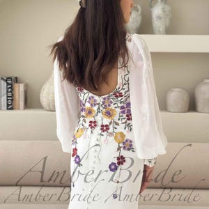 May include: White wedding dress with floral embroidery in yellow, purple, and red. The dress features long, sheer sleeves and a button-down back. The text "Amber Bride" is visible at the bottom of the image.