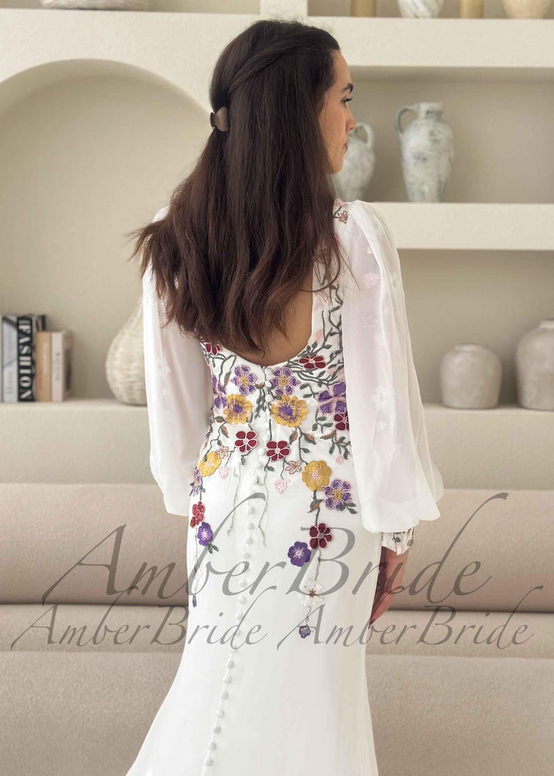 May include: White wedding dress with floral embroidery in yellow, purple, and red. The dress features long, sheer sleeves and a button-down back. The text "Amber Bride" is visible at the bottom of the image.