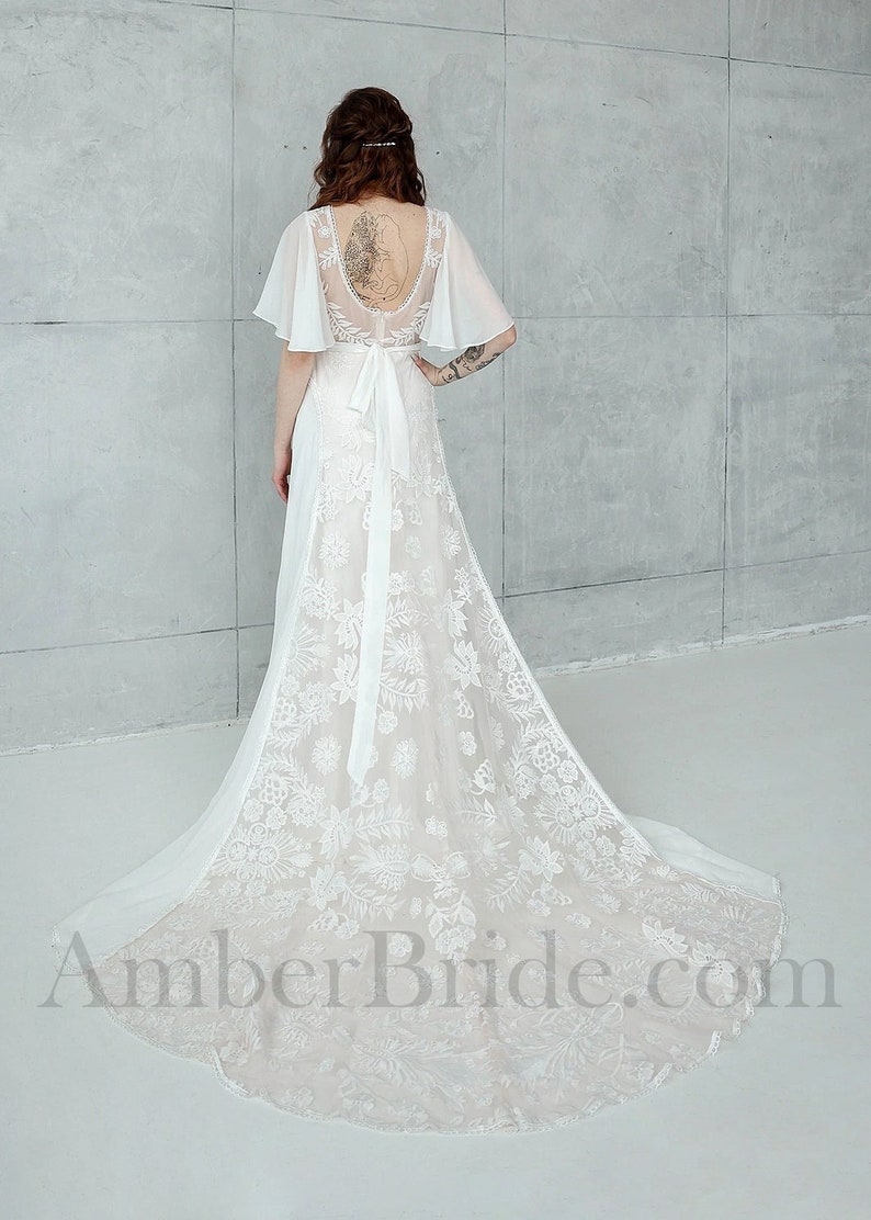 May include: A white lace wedding dress with a low back and a flowing skirt. The dress has a fitted bodice and a sash tied at the waist. The dress is shown from the back.