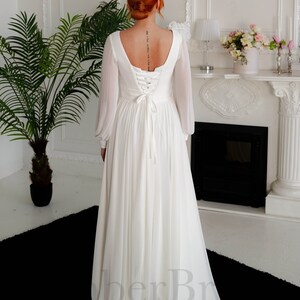 May include: A white chiffon wedding dress with long sleeves and a lace-up back. The dress has a scoop neckline and a flowing skirt.