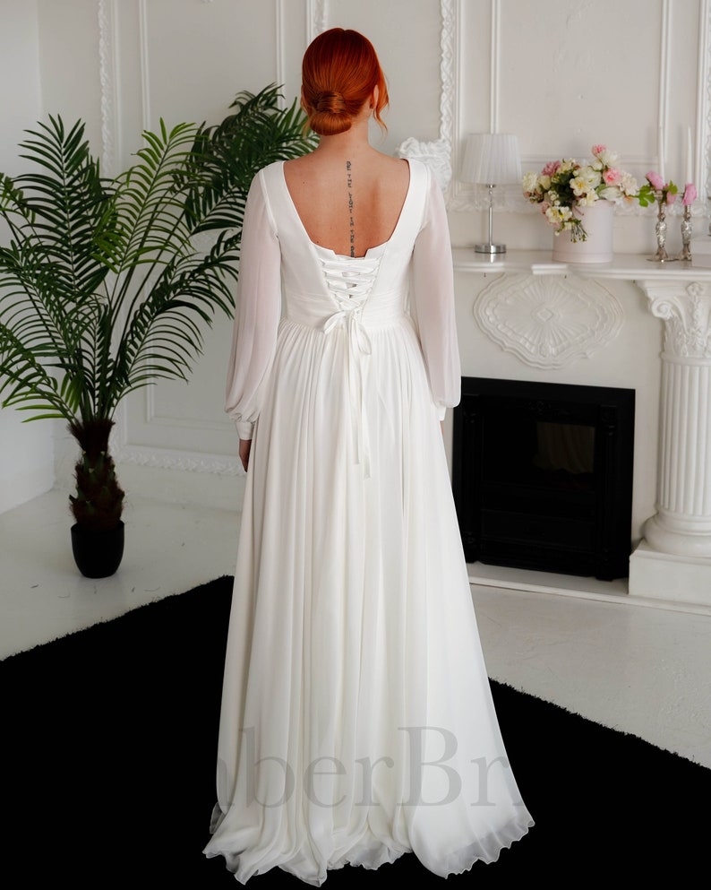 May include: A white chiffon wedding dress with long sleeves and a lace-up back. The dress has a scoop neckline and a flowing skirt.