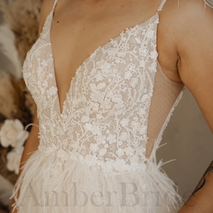 May include: A white wedding dress with a plunging neckline and a sheer bodice. The dress is adorned with delicate lace and sequins, and features a feather trim at the hem.