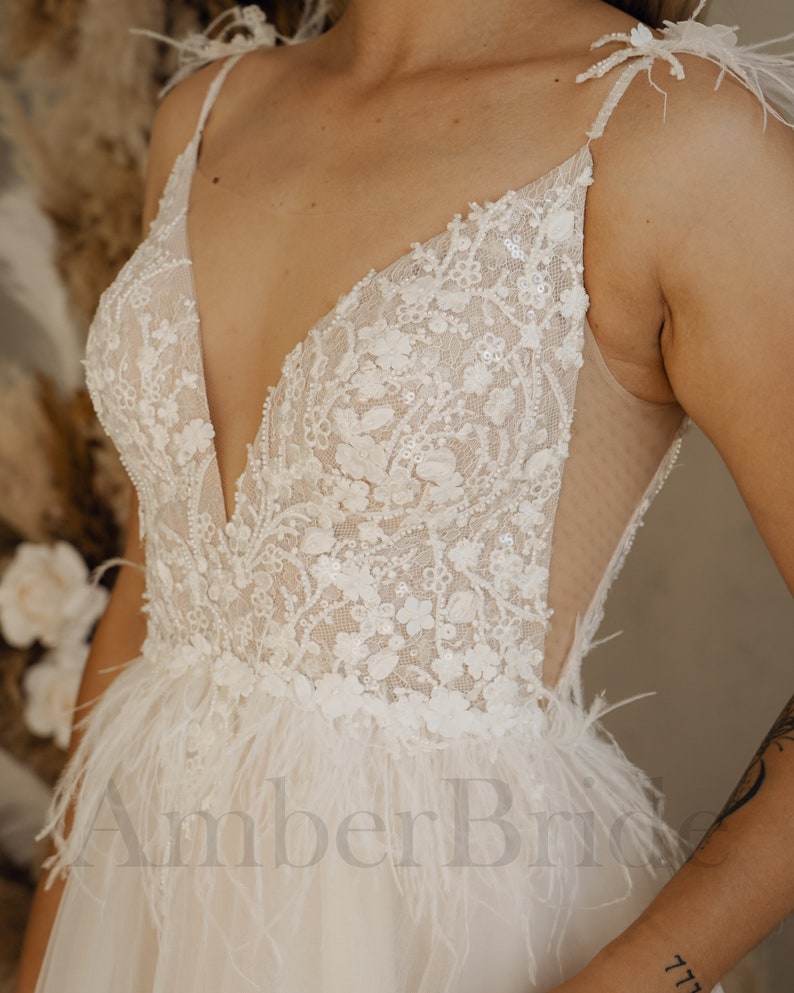 May include: A white wedding dress with a plunging neckline and a sheer bodice. The dress is adorned with delicate lace and sequins, and features a feather trim at the hem.