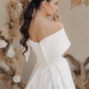 May include: A white off-the-shoulder wedding dress with a lace-up back and a flowing skirt. The dress has a simple, elegant design and is perfect for a modern bride.
