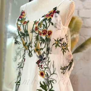 May include: A white wedding dress with a floral embroidery design. The embroidery features green leaves, red and yellow flowers, and gray accents.