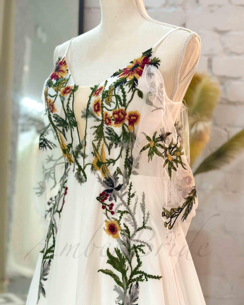 May include: A white wedding dress with a floral embroidery design. The embroidery features green leaves, red and yellow flowers, and gray accents.