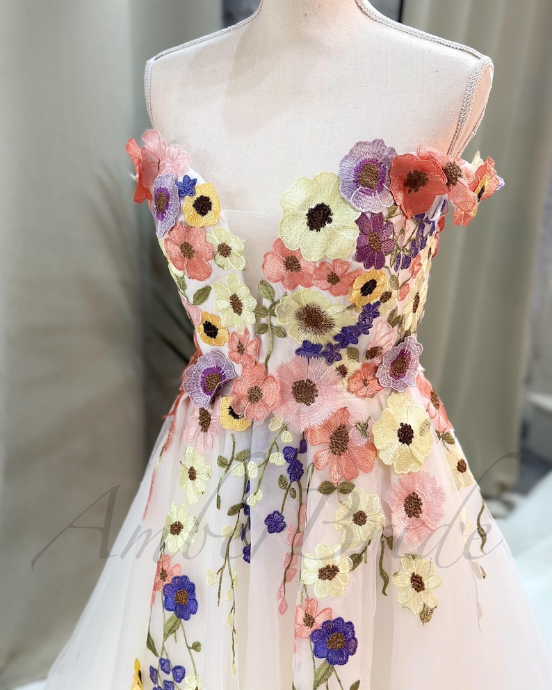 May include: A white off-the-shoulder dress with a floral embroidery design. The embroidery features a variety of colors, including pink, yellow, purple, and blue.