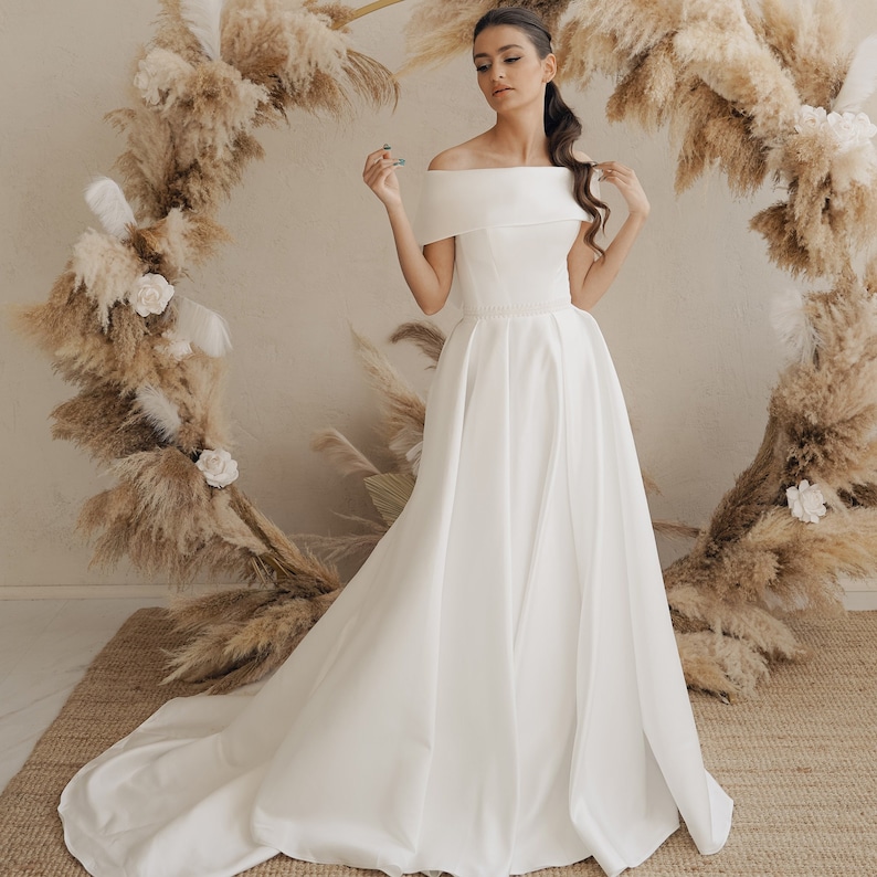 May include: A white off-the-shoulder wedding dress with a fitted bodice and a flowing A-line skirt. The dress is made of a smooth, satin-like fabric.
